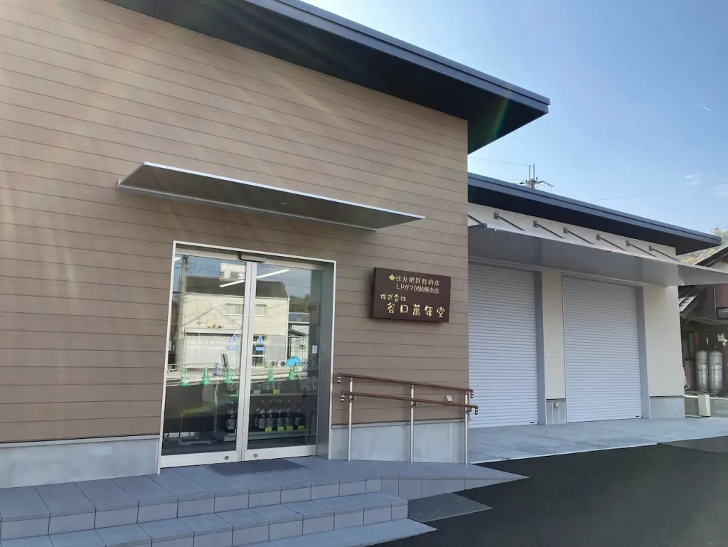 株式会社谷口萬年堂の本社建物外観。ベージュの外壁にガラス張りのエントランス、LPガス販売取扱所併設、バリアフリー対応のスロープ付き現代的な社屋