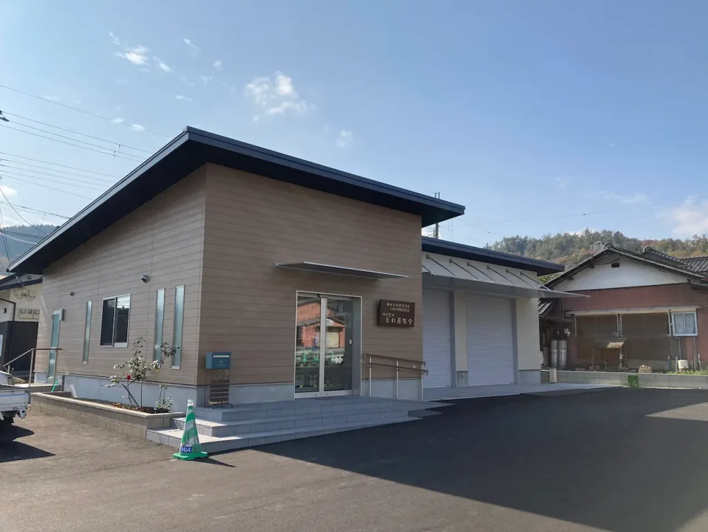 株式会社谷口萬年堂の本社建物全景。ベージュの横張り外壁と黒い屋根の現代的な平屋建て、LPガス販売取扱所併設、広い駐車場、背景に山並みが見える地域に根ざした事業所