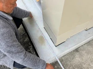リフォーム施工中の計測作業。グレーのフリースを着た職人が、コンクリート基礎の上に設置されたガス器具の寸法をメジャーで正確に測定。丁寧な施工作業の一場面