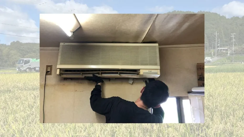 建設・リフォーム事業の様子。中央にエアコン設置作業を行う職人、背景に田園風景と配送トラック。地域密着型の建設サービスを表現した事業紹介画像