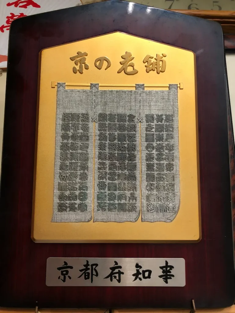 京都府知事による『京の老舗』認定証。暖簾デザインの金色の額装、伝統ある老舗企業としての公式認定を示す表彰額