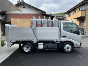 LPガス配送用小型トラック側面写真。銀色の荷台に5本の大型プロパンガスボンベ（LPガス表記）を積載、住宅街の道路脇に停車。背景に日本家屋と住宅が見える地域密着型配送サービスの車両